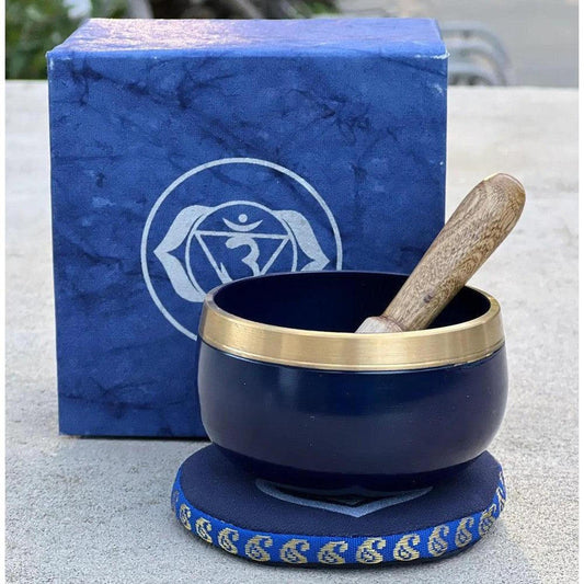 Midnight blue singing bowl with brass rim, wooden striker, and matching Ajna chakra gift box on a blue fabric cushion.