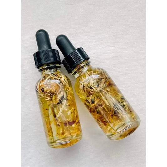 Two glass dropper bottles of golden Aura Cleanse ritual oil infused with dried botanicals and herbs.