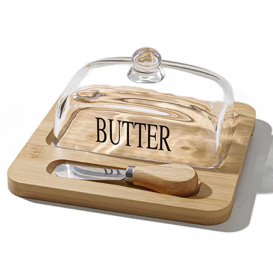 Light brown bamboo butter dish with a clear glass lid, labeled cover, and matching stainless steel butter spreader.