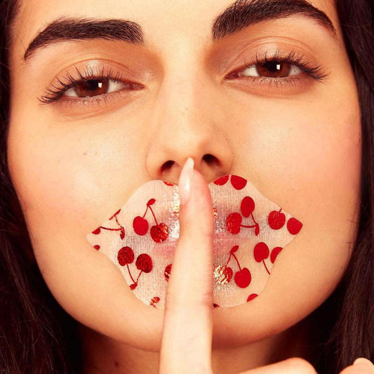Woman wearing a translucent hydrogel lip mask with a red cherry pattern, holding a finger to her lips in a hush gesture.