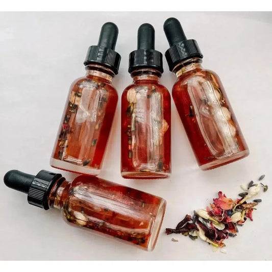 Four glass dropper bottles filled with red ritual oil, rose petals, and jasmine blossoms on a white background.