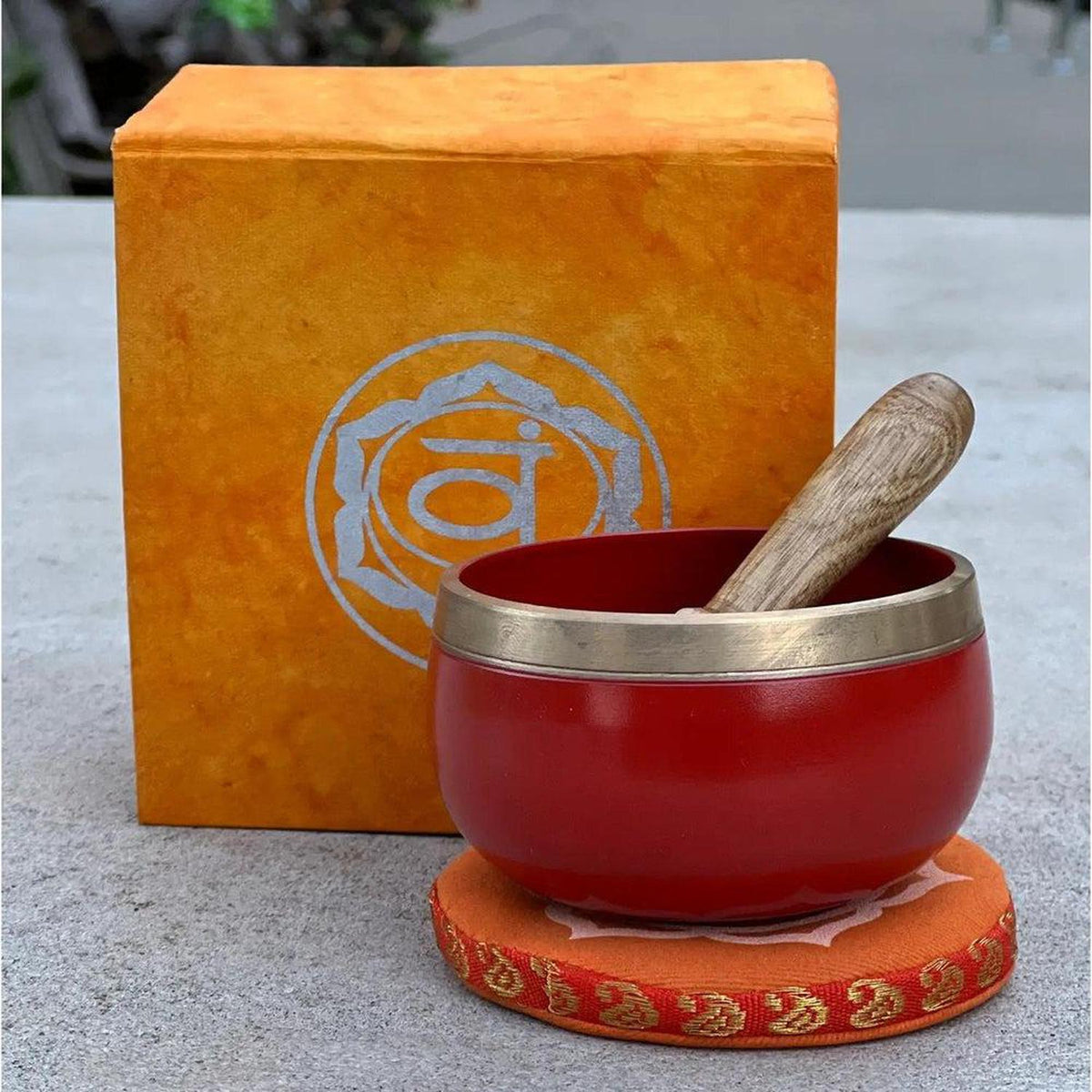 Red brass singing bowl with wooden mallet, sitting on an orange cushion in front of a matching Sacral Chakra gift box.