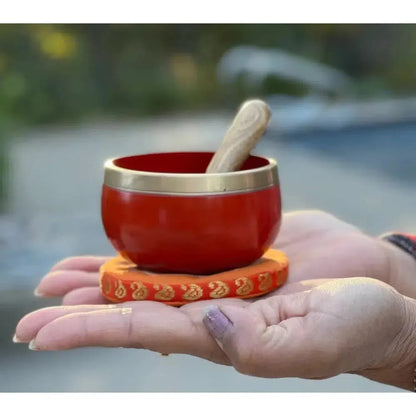 Small red brass Sacral Chakra singing bowl with a wooden mallet, resting on an orange embroidered cushion in a palm.