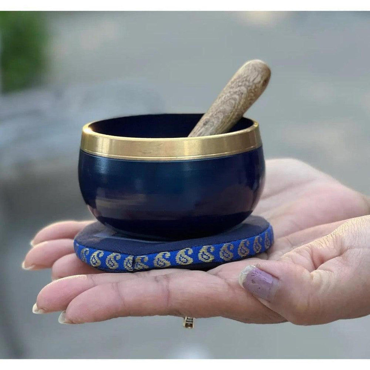 Indigo blue singing bowl with a polished brass rim, held in a hand with a wooden mallet and matching fabric cushion.